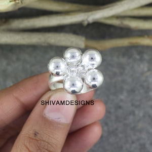 May include: A silver ring featuring a flower design with six round, polished spheres. The ring is held by a hand, with the brand name "SHIVAMDESIGNS" visible. The ring is a piece of jewelry.