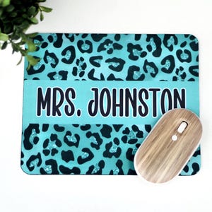 May include: A personalized mousepad with a teal leopard print design and the name "MRS. JOHNSTON" in bold, black letters. A wooden wireless mouse rests on the pad. The mousepad is rectangular.