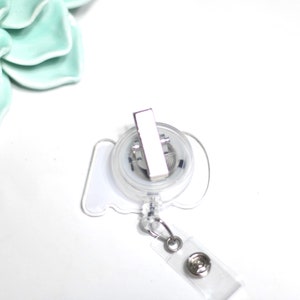 Pode incluir: Um porta-crach&aacute; retr&aacute;til transparente com um clipe de metal prateado e uma al&ccedil;a de pl&aacute;stico transparente. O carretel tem uma forma branca estilizada. O fundo &eacute; branco com uma suculenta de cer&acirc;mica verde claro.