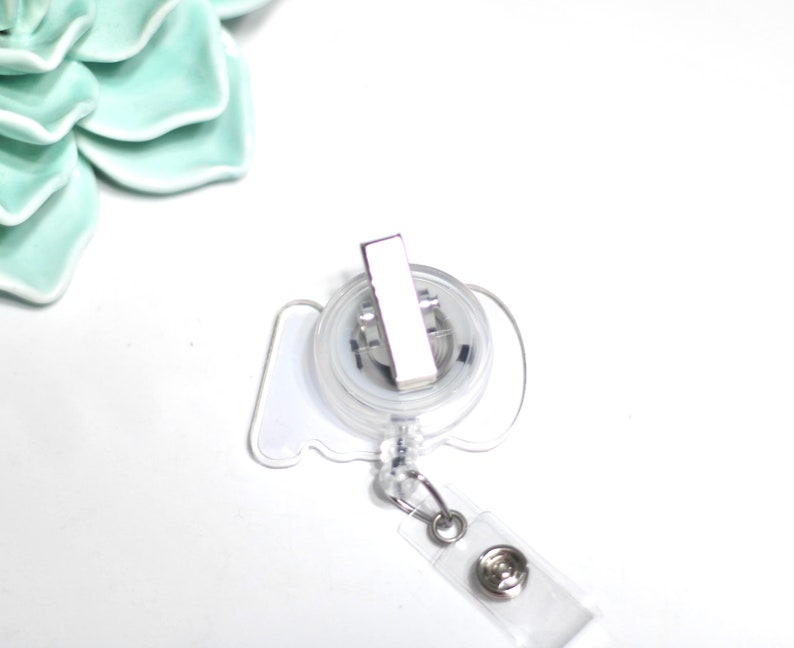 Pode incluir: Um porta-crach&aacute; retr&aacute;til transparente com um clipe de metal prateado e uma al&ccedil;a de pl&aacute;stico transparente. O carretel tem uma forma branca estilizada. O fundo &eacute; branco com uma suculenta de cer&acirc;mica verde claro.