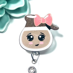 May include: A brown and white cartoon coffee cup with a pink bow and glitter. The cup has a smiling face with big eyes. The cup is attached to a white retractable badge holder.