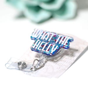 May include: A badge reel with the phrase "WHAT THE HELL" in white lettering. The badge reel has a clear plastic retractable cord and a metal clip. The badge itself is blue with red and white glitter accents.