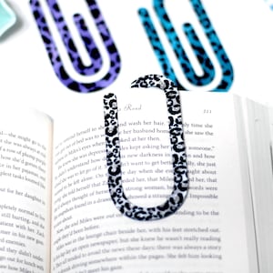 May include: A black and white leopard print bookmark in the shape of a paperclip is placed on the pages of a book. Two other leopard print bookmarks in purple and teal are in the background. The book is open to page 211.
