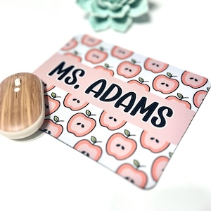 May include: A personalized mousepad with a pink apple pattern and the text "MS. ADAMS" in black. A wooden-look wireless mouse is next to the mousepad. The background is white.