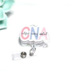 Pode incluir: Um crach&aacute; retr&aacute;til transparente de CNA com as palavras "CNA certified nursing assistant" em letras rosa e azul. O crach&aacute; tem um clipe de pl&aacute;stico transparente e um cord&atilde;o retr&aacute;til com um fecho de metal.