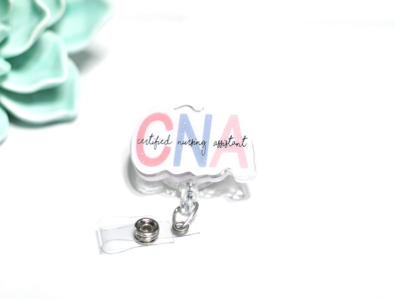 Pode incluir: Um crach&aacute; retr&aacute;til transparente de CNA com as palavras "CNA certified nursing assistant" em letras rosa e azul. O crach&aacute; tem um clipe de pl&aacute;stico transparente e um cord&atilde;o retr&aacute;til com um fecho de metal.