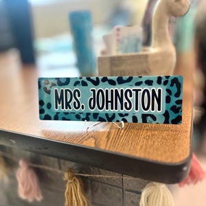 May include: A rectangular desk sign with the text "MRS. JOHNSTON" in white letters on a teal leopard print background. The sign is displayed on a wooden desk with a tassel garland.