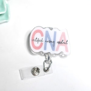 Pode incluir: Um porta-crach&aacute; CNA transparente com um design branco, rosa e azul. O porta-crach&aacute; tem o texto "CNA certified nursing assistant" em uma fonte de script. O porta-crach&aacute; tem um clipe de pl&aacute;stico transparente e um fecho de metal prateado.