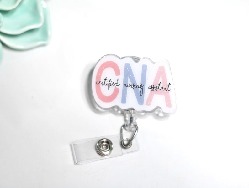 Pode incluir: Um porta-crach&aacute; CNA transparente com um design branco, rosa e azul. O porta-crach&aacute; tem o texto "CNA certified nursing assistant" em uma fonte de script. O porta-crach&aacute; tem um clipe de pl&aacute;stico transparente e um fecho de metal prateado.