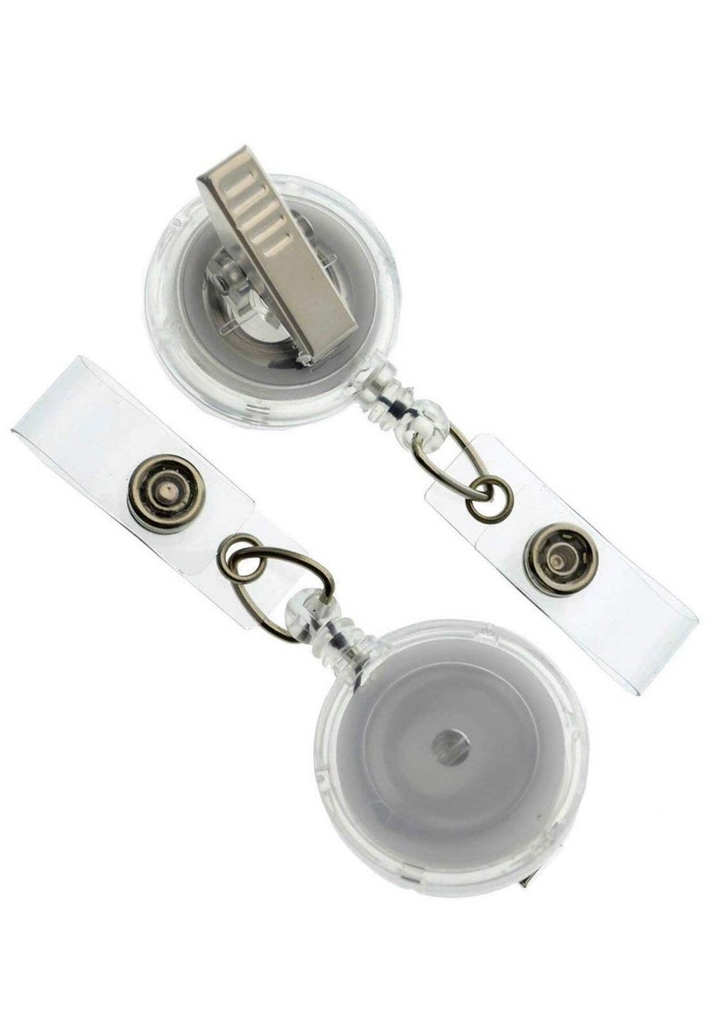 Pode incluir: Dois porta-crach&aacute;s retr&aacute;teis de pl&aacute;stico transparente com clipes de metal prateado e tiras de vinil transparente. Os carret&eacute;is de crach&aacute; t&ecirc;m um design circular e s&atilde;o conectados &agrave;s tiras com an&eacute;is de metal.