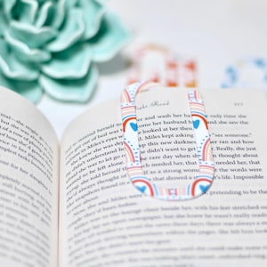 May include: A colorful bookmark with a rainbow and heart design is placed on an open book. The bookmark has a U-shape and is decorated with red, blue, and white patterns. The book's pages are filled with text.