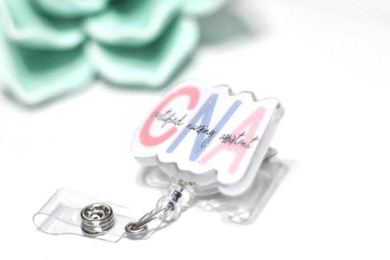 Pode incluir: Um porta-crach&aacute; branco com uma al&ccedil;a de pl&aacute;stico transparente e um fecho de press&atilde;o de metal. O crach&aacute; apresenta as letras "CNA" em rosa e azul, com as palavras "certified nursing assistant" em script. Uma suculenta verde claro no fundo.