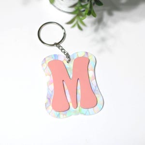 May include: A pink letter "M" keychain with a silver keyring and chain. The letter is set against a background of iridescent, pastel-colored shapes. The keychain is a decorative accessory.