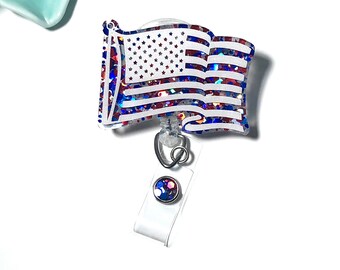 Pode incluir: Um porta-crachá de plástico transparente com um design de bandeira americana de glitter vermelho, branco e azul. O porta-crachá tem um carretel retrátil de plástico transparente e um botão redondo iridescente.