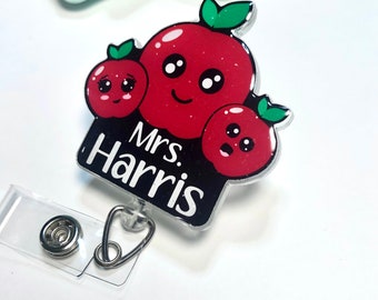 Pode incluir: Um crachá de identificação de professor preto e branco com o texto "Mrs. Harris" e um design de maçã vermelha com três maçãs de desenho animado com rostos.