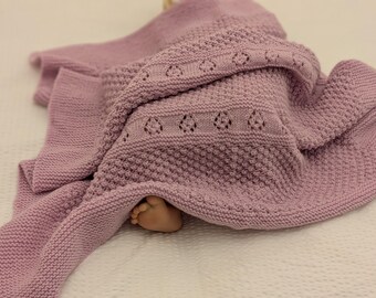May include: A soft, dusty lavender knitted blanket featuring a textured seed stitch pattern and a horizontal band of delicate eyelet lace detailing. The fabric is draped in folds, revealing a small foot peeking out from underneath the knit.
