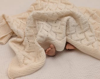 May include: A cream-colored knitted blanket featuring a textured basketweave pattern with a garter stitch border. The fabric is draped over two small feet, showcasing the intricate cable-like knit design and the soft, off-white yarn construction.