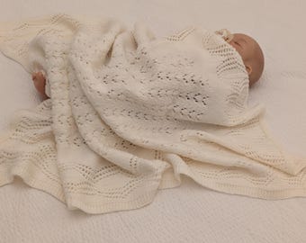 May include: A cream-colored knitted baby blanket featuring an intricate openwork lace pattern with a scalloped edge. The soft, lightweight textile is draped over a doll, showcasing the detailed texture and delicate weave of the fabric.