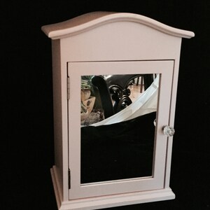 May include: A small, pale pink wooden cabinet with a mirrored door and a crystal knob. The cabinet has a slightly arched top and sits on a black surface.