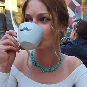 Op de afbeelding: Een persoon drinkt uit een witte koffiemok met een zwart snorontwerp. Ze draagt een witte top en een turquoise ketting. De achtergrond toont een buitencaf&eacute; met andere mensen.