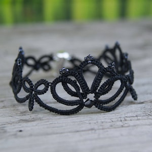 Op de afbeelding: Zwarte kanten choker ketting met een bloemenmotief. Het ingewikkelde patroon heeft lussen en kralenaccenten. De ketting is vastgezet met een zilverkleurige sluiting. De achtergrond is een vage houten ondergrond.