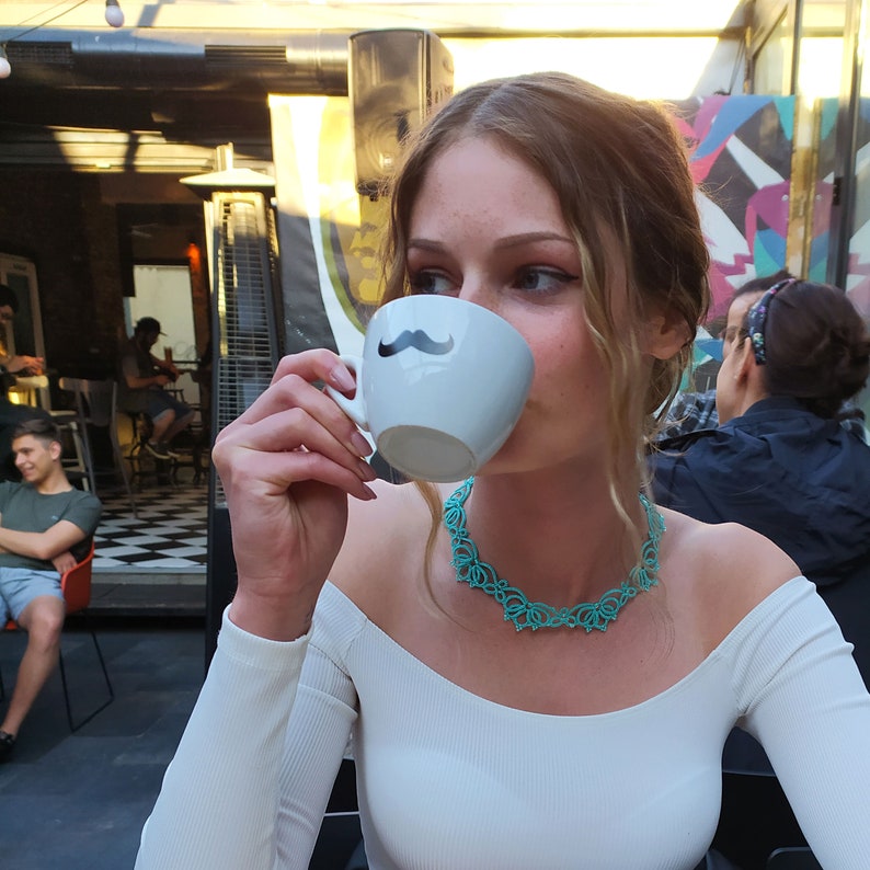 Op de afbeelding: Een persoon drinkt uit een witte koffiemok met een zwart snorontwerp. Ze draagt een witte top en een turquoise ketting. De achtergrond toont een buitencaf&eacute; met andere mensen.