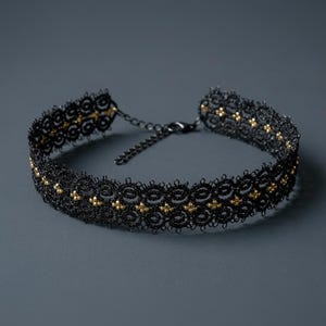 May include: A black lace choker necklace with gold-colored bead accents. The necklace features a delicate, intricate design and a chain clasp for adjustable sizing. The choker is displayed against a dark gray background.