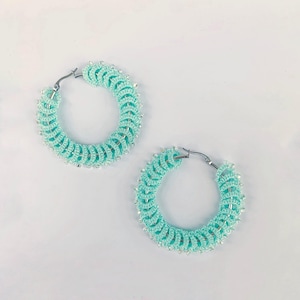 Op de afbeelding: Een paar turquoise oorringen. De oorbellen zijn gehaakt met een spiraalvormig ontwerp en geaccentueerd met kleine, zilverkleurige kralen. De oorringen hebben een zilverkleurige sluiting.