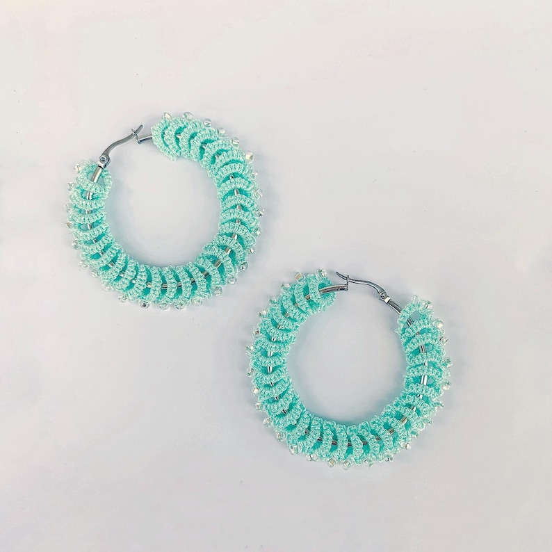 Op de afbeelding: Een paar turquoise oorringen. De oorbellen zijn gehaakt met een spiraalvormig ontwerp en geaccentueerd met kleine, zilverkleurige kralen. De oorringen hebben een zilverkleurige sluiting.