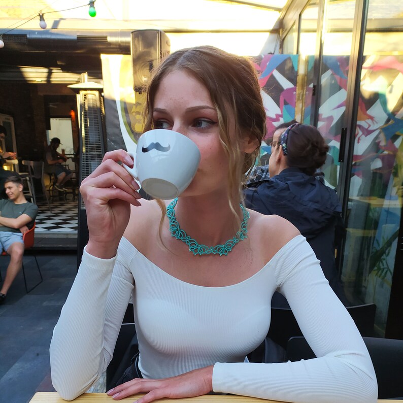 Op de afbeelding: Een witte keramische mok met een zwart snorontwerp wordt naar de mond gebracht. Een turkooizen ketting met een bloemenontwerp wordt om de nek gedragen. De persoon draagt een witte off-the-shoulder longsleeve.