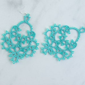 Op de afbeelding: Een paar turquoise kanten oorbellen met zilverkleurige haken. De oorbellen hebben een ingewikkeld, open ontwerp met een bloemenpatroon. De oorbellen worden gepresenteerd op een wit en grijs marmeren oppervlak.