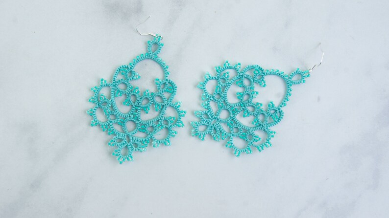 Op de afbeelding: Een paar turquoise kanten oorbellen met zilverkleurige haken. De oorbellen hebben een ingewikkeld, open ontwerp met een bloemenpatroon. De oorbellen worden gepresenteerd op een wit en grijs marmeren oppervlak.