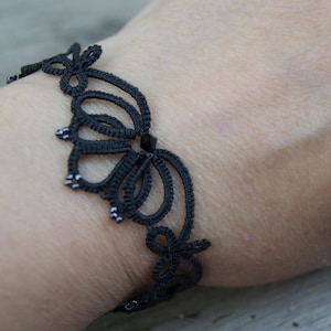 Op de afbeelding: Zwarte kanten armband met een bloemmotief. De armband is gemaakt van een delicaat, geweven materiaal en heeft kleine, zilverkleurige kralen. De armband wordt om de pols gedragen.
