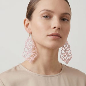 Op de afbeelding: Zachtroze, open oorbellen met een delicaat, wervelend ontwerp. De oorbellen zijn druppelvormig en hangen aan goudkleurige haken. De oorbellen zijn een statement piece en voegen een vleugje elegantie toe.