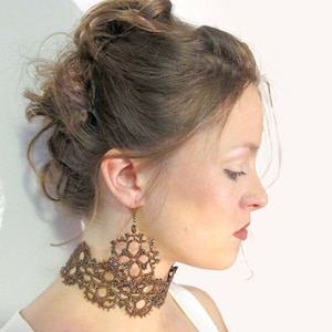 Może przedstawiać: Kobieta nosi brązowy naszyjnik typu choker z koronki i pasujące kolczyki. Kolczyki są duże i bogato zdobione, z misternym, ażurowym wzorem. Biżuteria ma ciepły, ziemisty brązowy kolor, a kobieta ma spięte włosy.