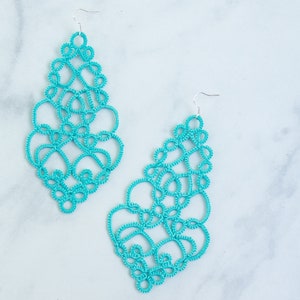 Op de afbeelding: Twee turquoise, opengewerkte oorbellen in druppelvorm worden getoond tegen een witte marmeren achtergrond. De oorbellen hebben een delicaat, kantachtig ontwerp met zilveren haken voor het oor.