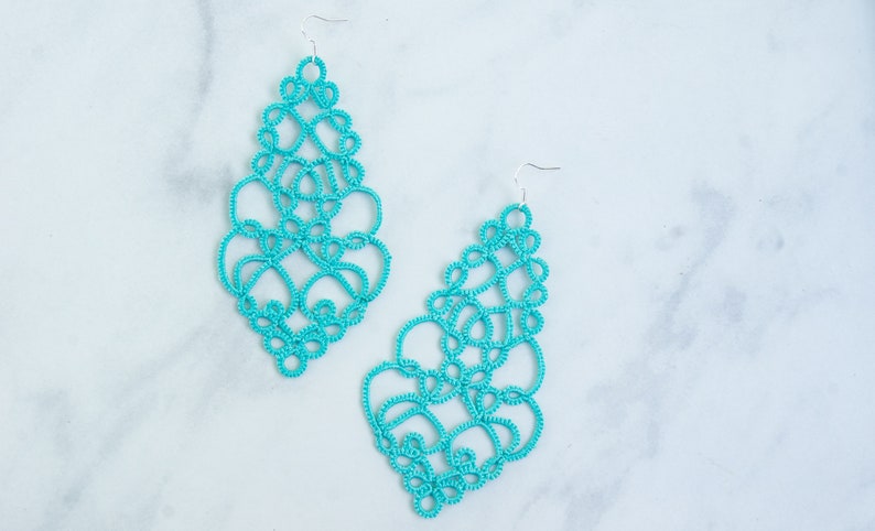 Op de afbeelding: Twee turquoise, opengewerkte oorbellen in druppelvorm worden getoond tegen een witte marmeren achtergrond. De oorbellen hebben een delicaat, kantachtig ontwerp met zilveren haken voor het oor.