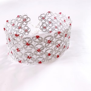 Può includere: Un braccialetto di perline argentate e rosse con un design delicato e intricato. Il braccialetto è realizzato con piccole perline argentate che sono intrecciate insieme per creare uno schema di fiori. I fiori sono accentuati da piccole perline rosse.