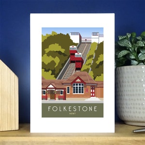 May include: An illustration of the Folkestone Funicular, a historic cable railway in Folkestone, Kent. The illustration shows the red cable cars ascending the hill, with a green building at the bottom of the hill.