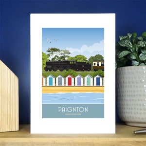 Peut inclure: Une illustration colorée d'un train voyageant le long d'une plage avec des cabines de plage au premier plan. Le texte "Paignton South Devon" est en bas de l'image.