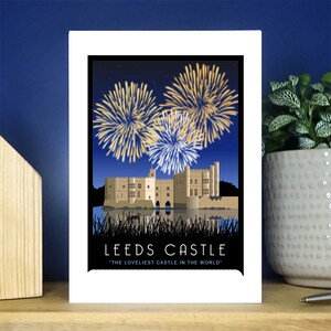 May include: A white framed print with a black border featuring a colourful illustration of Leeds Castle with fireworks exploding in the night sky. The text "Leeds Castle" and "The loveliest castle in the world" is printed below the image.