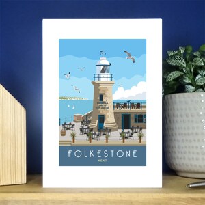 May include: A colourful illustration of a lighthouse with a blue sky and sea in the background. The lighthouse is on a pier with tables and chairs. The text "FOLKESTONE KENT" is at the bottom of the image.