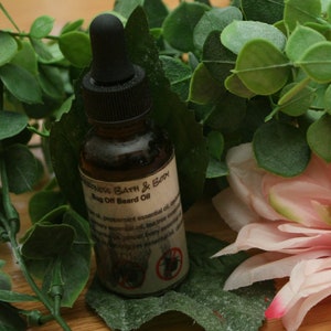 May include: A small brown glass bottle with a dropper top and a label that reads "Wilderness Bath & Body Bug Off Beard Oil". The bottle is surrounded by green leaves and a pink flower.