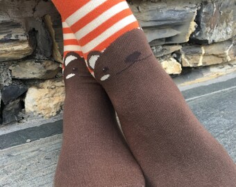 May include: Brown socks with orange and white striped cuffs and a brown bear face design on the top of the sock.