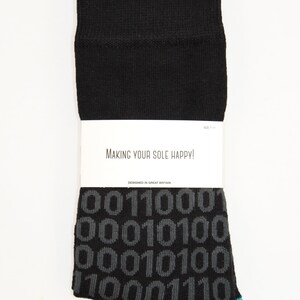 Op de afbeelding: Zwarte sokken met een blauwe rand en een grijs binair codepatroon. De sokken hebben een wit label met de tekst "Making your sole happy!" en "Designed in Great Britain".