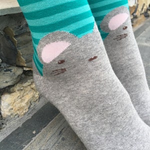 May include: A pair of grey socks with a teal and white striped pattern. Each sock features a grey mouse face with pink ears and brown eyes.