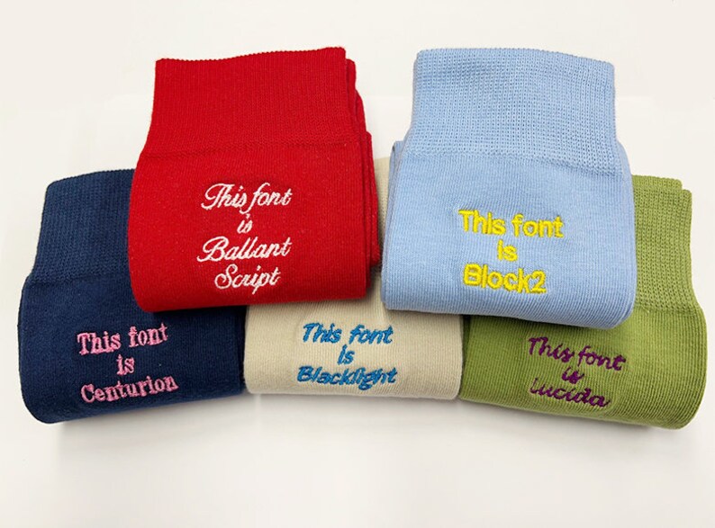 Op de afbeelding: Zes paar sokken van verschillende stoffen met geborduurde tekst. De tekst op de sokken luidt "This font is Centurion", "This font is Ballant Script", "This font is Blacklight", "This font is Block2", "This font is Lucida".