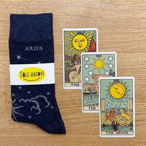 Puede incluir: Un par de calcetines de color gris oscuro con un patrón de estrellas blancas y las palabras "ARIES" y "SOLE HAPPY!" impresas en ellos. También se muestran tres cartas del tarot: El Sol, La Estrella y La Luna.