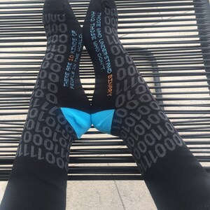 Op de afbeelding: Een paar zwarte sokken met een grijs binair codepatroon. De sokken hebben een blauwe en turquoise accent bovenaan. De tekst op de sokken luidt "Er zijn 10 soorten mensen in de wereld. Degenen die binair begrijpen en degenen die dat niet doen."