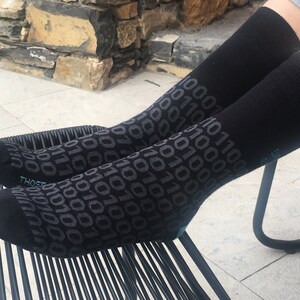 Op de afbeelding: Een paar zwarte sokken met een herhalend patroon van grijze 1s en 0s. De sokken liggen op een zwarte stoel.